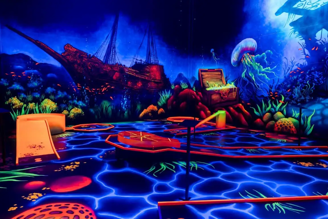 Unterwasserwelt der Minigolfanlage: ein leuchtendes Schiffswrack, eine Qualle und eine Schatztruhe in Schwarzlichtfarben, davor die Bahnen.